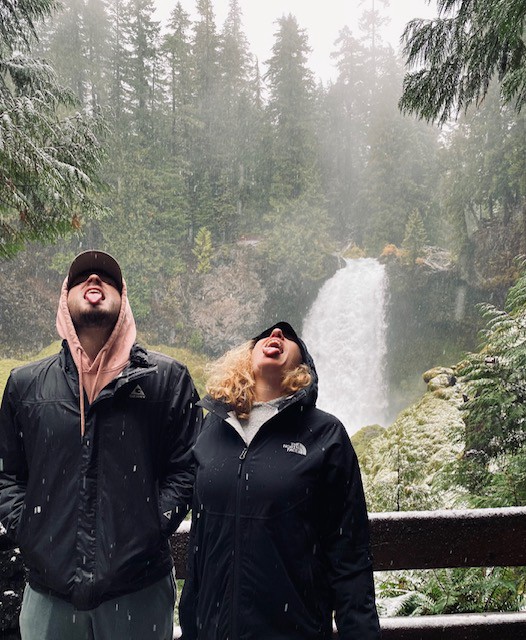 A young man and his mother in black raincoats, stand in front of a waterfall with their mouths open to catch snowflakes on their tongues